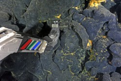 ROPOS’s manipulator reaches to take a sample of glassy, fresh basalt from the 2015 eruption. Orange bacterial mats coat the crevices. (Credit: NSF-OOI/UW/ROPOS; V15)