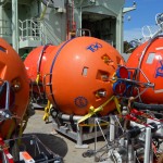 Sub-surface flotation spheres and controllers for the two Flanking Moorings and the Global Profiler Mooring sit on deck awaiting deployment. ADCPs (red circles with blue rims) can be seen on the top of two of the Flanking Mooring spheres. These instruments will measure water velocity from the depth of the spheres (500 m) to the sea surface. The 64” spheres in the background provide floatation at the top of the moorings. The upper buoyancy on the flanking moorings is deployed close to the surface, at about 2o to 30 m depth; and at the Irminger Sea site the two flanking moorings each carry 4 pairs temperature/conductivity and velocity instruments at depths matching those of the OSNAP array. (Photo Credit: Sheri N. White)