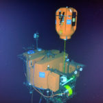 Resting 200 meters under water at the base of the Axial Seamount, the Cabled Shallow Profiler Mooring science pod hovers above its docking station.  Soon it will begin its ascent toward the surface, sampling the water column using the instruments (black cylinders) attached to the pod. Credit: NSF-OOI/UW/ISS; Dive R1842; V15