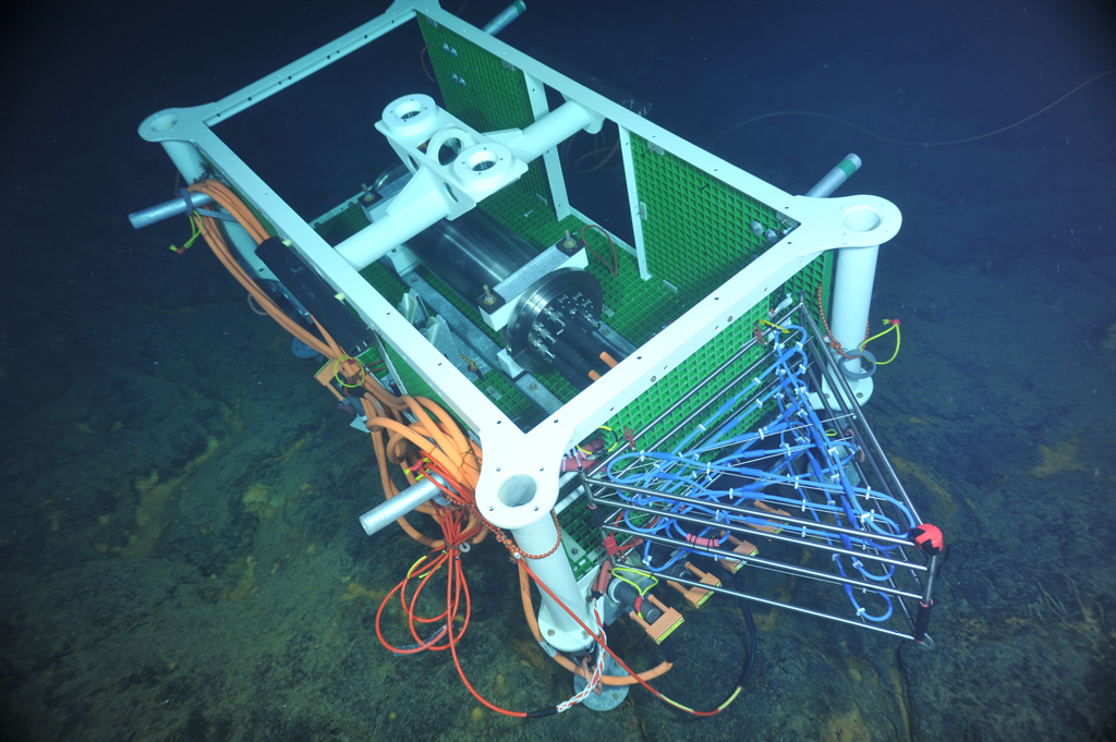 Medium-power junction box installed in the ASHES hydrothermal field of Axial Seamount’s caldera. The titanium cylinder inside the node frame hosts power converters, data ports, and shore-linked communication capabilities. This J-box also hosts a cabled 3D thermistor array (the triangular-shape frame with blue cables) that was deployed for testing during the expedition. (Photo credit: NSF-OOI/UW/CSSF)