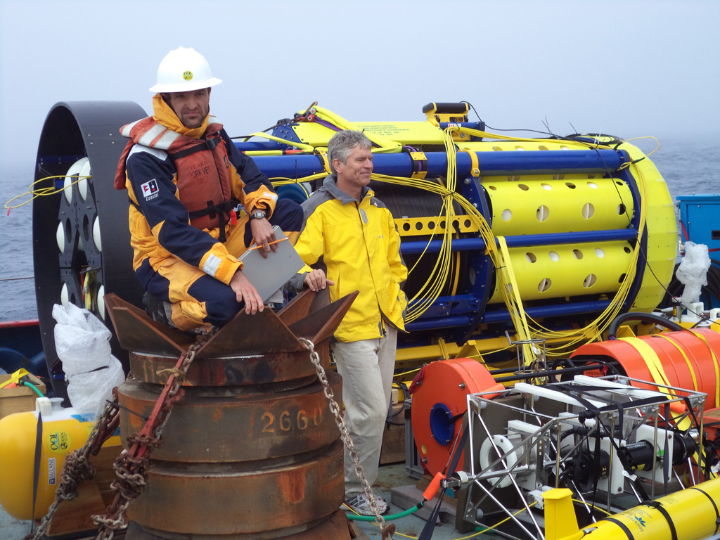 SIO scientist Matthias Lankhorst performs deployment Quality Control (QC) while perched on a mooring anchor with Chief Scientist Uwe Send next to the global surface piercing profiler. Note glider in foreground and McLane profiler on left. (Photo Credit: Bill Bergen, OOI Program Management Office)